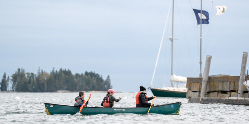 The image shows three people in a canoe on a lake. There is a sailboat and a dock in the background. The sky is overcast. The people are wearing life jackets and appear to be enjoying a day out on the water. The overall scene is peaceful and serene.